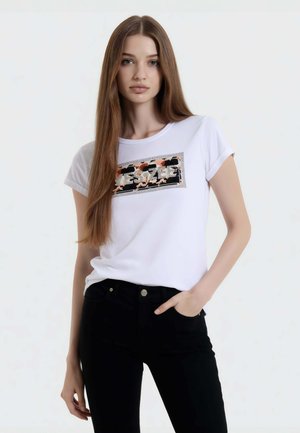 Giovane donna con capelli lunghi e lisci che indossa una t-shirt bianca con grafica e jeans neri, una mano in tasca, su uno sfondo bianco semplice.