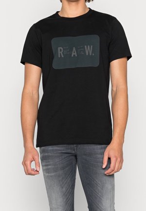 Mężczyzna ubrany w czarną, krótki rękaw koszulkę z ciemnym prostokątnym logo o treści "R A W.", oraz szare jeansy, stoi na tle jednolitego tła.