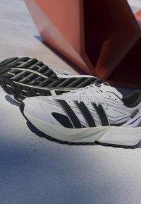 White athletic shoes with black accents and textured outsole, featuring a sleek design, laces, and a visible brand label on the side.