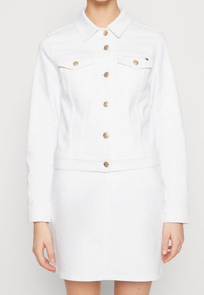 Mujer con una chaqueta de mezclilla blanca abotonada con botones dorados y una falda blanca a juego, de pie contra un fondo liso.
