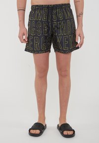 Pantaloni da bagno neri con un audace motivo testuale giallo, fascia in vita elastica e vestibilità rilassata; indossati con sandali neri senza chiusura.