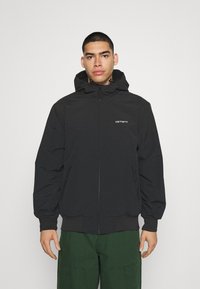 Veste zippée noire avec capuche, présentant des poignets et une taille élastiques, logo sur la poitrine, et deux poches latérales. Texture lisse.