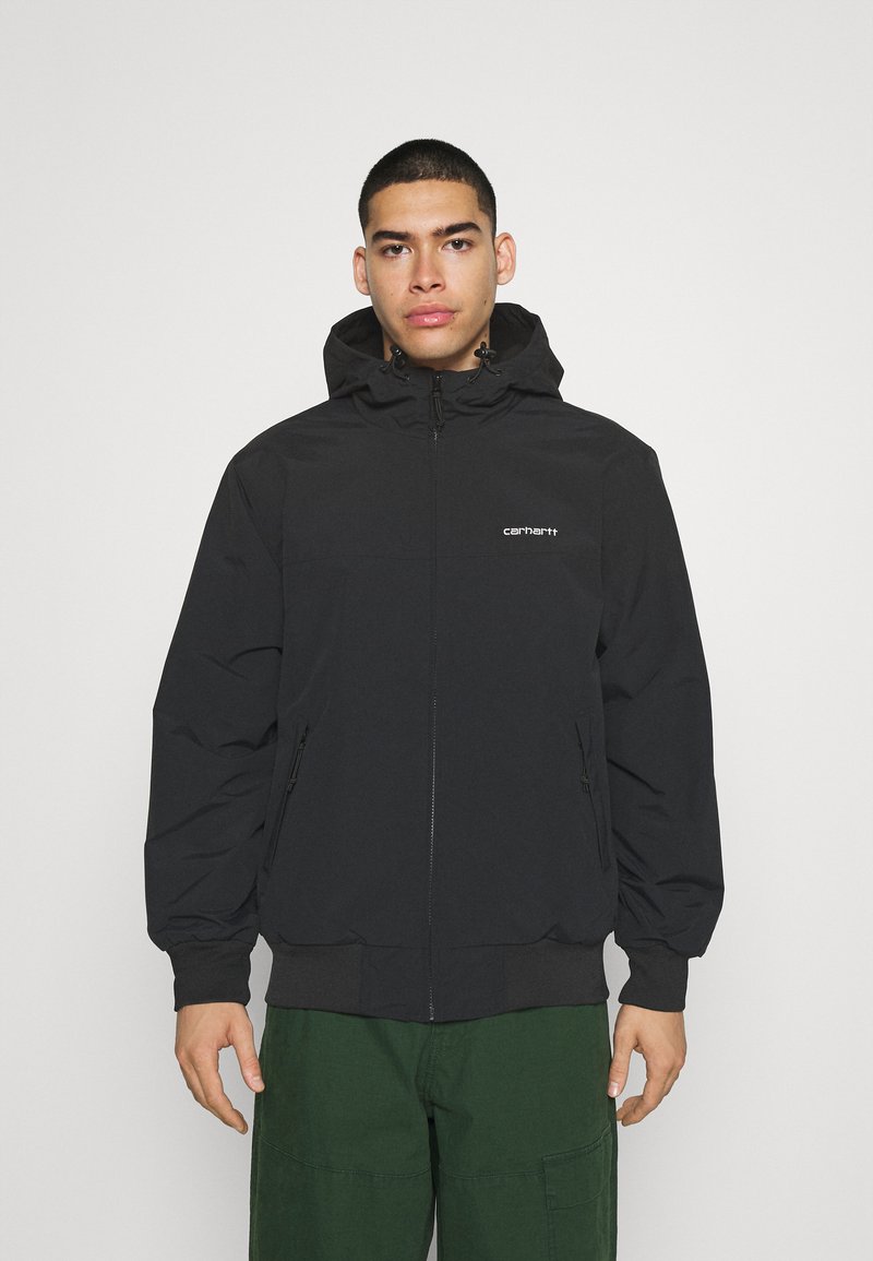 Veste zippée noire avec capuche, présentant des poignets et une taille élastiques, logo sur la poitrine, et deux poches latérales. Texture lisse.