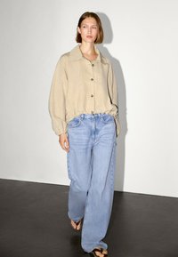 Camicia button-up beige chiaro con un ampio colletto; maniche a sbuffo. Jeans blu a vestibilità comoda. Indossato con ciabatte nere su un pavimento nero.