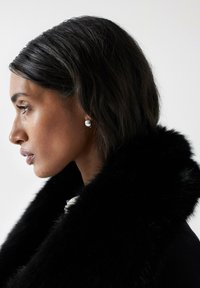 Un col en fourrure noire s'enroule autour du cou, mettant en valeur une boucle d'oreille en argent. Des cheveux lisses et sombres tombent doucement contre la peau.