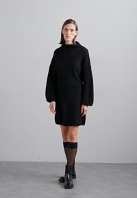 Rochie tricotată neagră cu gât înalt și mâneci tip balon, având o croială lejeră. Modelul poartă șosete negre până la genunchi și botine.