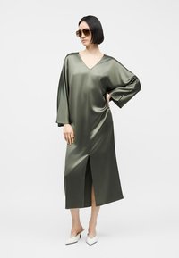 Γυναίκα με υπερμεγέθη σατινέ φόρεμα σε ελιά πράσινο χρώμα με V-λαιμόκοψη, μπροστινή σχισμή, μακριά φαρδιά μανίκια, γυαλιά ηλίου και λευκά μυτερά παπούτσια με τακούνι.