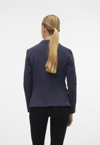 Navyblauer, glatter Stoffblazer mit tailliertem Schnitt, Ein-Knopf-Verschluss und Rückennähten; von hinten betrachtet, kombiniert mit schwarzen Hosen.
