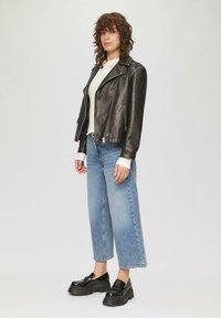 Schwarze Lederbikerjacke, leichter Pullover, weit geschnittene blaue Jeans und schwarze Plateaulöcher. Lockige Haare sichtbar, stehende Pose.