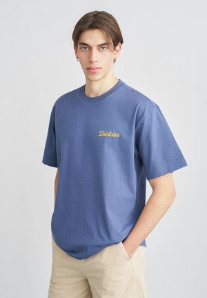 Joven con cabello corto castaño que lleva una camiseta azul de Dickies y pantalones beige, posando con una mano en el bolsillo contra un fondo blanco liso.