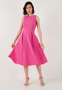 Fuchsia sleeveless dress with a fitted bodice, flared skirt, smooth texture, and knee-length hem. Model wears colorful strappy sandals.