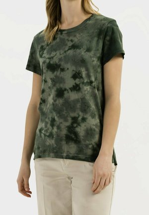 Femme portant un t-shirt à manches courtes tie-dye vert foncé et un pantalon beige clair, debout avec les bras détendus le long du corps.