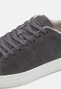 Zapatilla de suede gris con cordones trenzados grises, suela de goma blanca y un sutil detalle de costura a lo largo del borde. Diseño minimalista y líneas limpias.