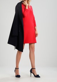 Robe rouge à mi-longueur avec des manches longues, associée à un manteau bleu marine drapé sur une épaule. Présente des chaussures à talons hauts bleu marine avec des brides autour de la cheville.