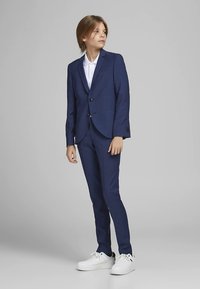 Tamsiai mėlynas kostiumas iš lygios medžiagos, pritaikytas kūno linijoms, su dviem mygtukais, derinamas su balta marškiniais ir baltais sportbačiais.