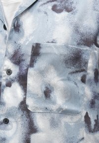 Camisa de botones azul claro con estampado floral abstracto en blanco y negro y un bolsillo frontal en el pecho, sobre una camiseta interior blanca.