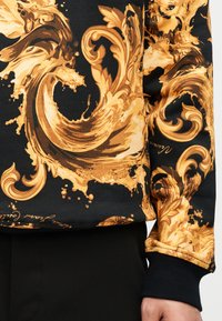 Μαύρο φούτερ με περίτεχνα, σπειροειδή μοτίβα σε χρυσό μπαρόκ στυλ και πιτσιλιές, φορεμένο με μαύρο παντελόνι, εμφανής η μανσέτα και το χέρι.