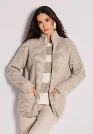 Frau in beigem, geripptem Reißverschluss-Cardigan über einem gestreiften, beige-weißen Pullover, kombiniert mit beigen Hosen und großen ovalen Ohrringen.