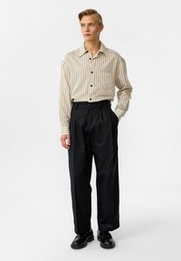 Lichtbeige gestreepte lange mouwen shirt met een zak, gecombineerd met hooggetailleerde zwarte broek en zwarte schoenen, tegen een witte achtergrond.
