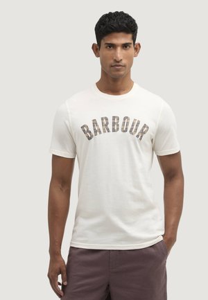 Mężczyzna w białej koszulce z krótkim rękawem z napisem "BARBOUR" w kratkę i ciemnych szortach, stojący na jednolitym jasnym tle.