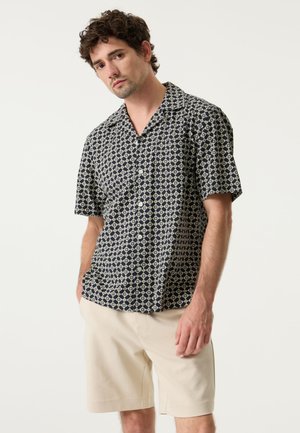 Camisa de manga corta con un patrón geométrico en azul marino y crema, con botones en la parte delantera, cuello y corte relajado. Combinada con pantalones cortos beige.