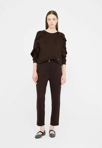 Brązowy sweter z falbankami na rękawach, zestawiony z brązowymi spodniami typu slim fit. Całość uzupełniają złote akcenty oraz płaskie buty o fakturowanym wzorze.