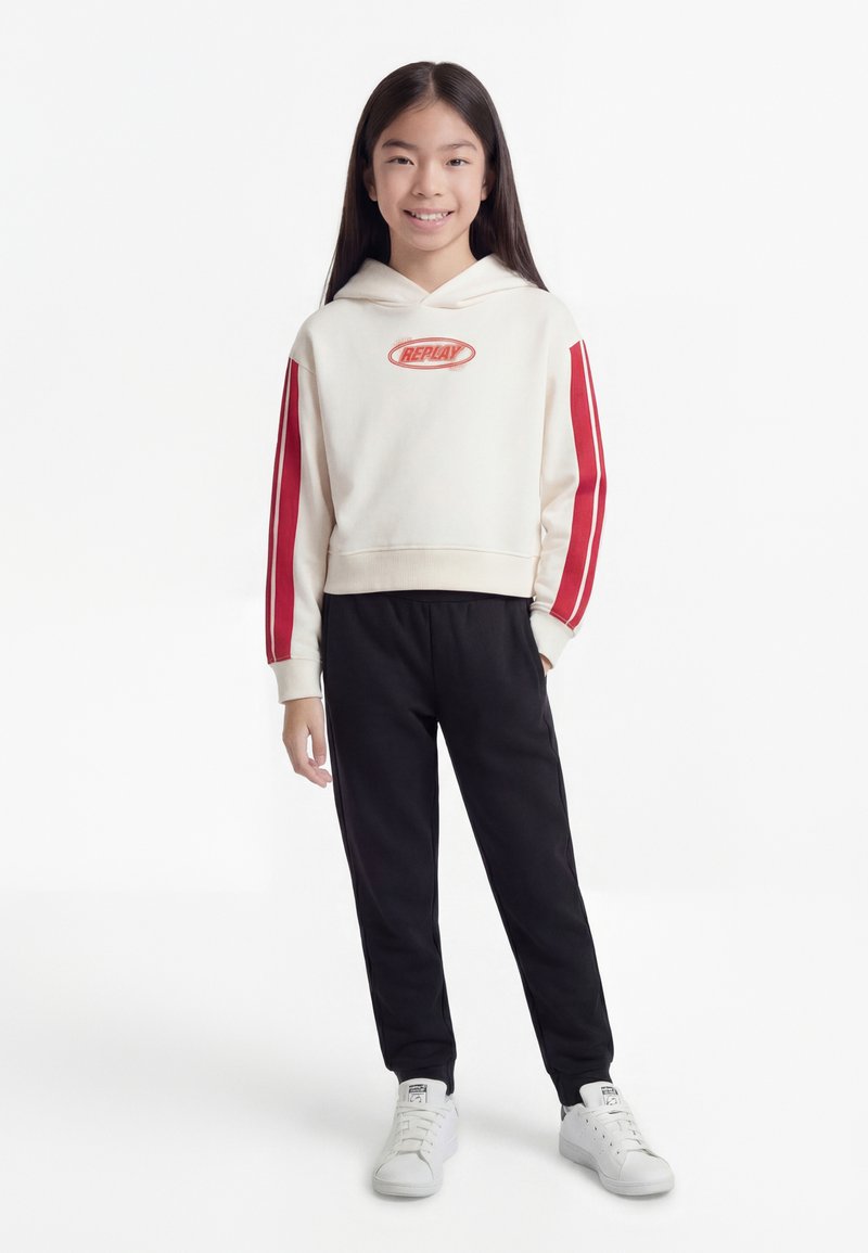 Jeune personne debout, portant un sweat à capuche blanc avec des rayures rouges et le logo "Replay", un pantalon noir et des baskets blanches, souriant à la caméra.