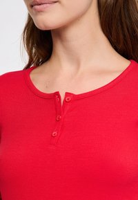 Czerwony, prążkowany top z długim rękawem, o okrągłym dekolcie i trzech zapięciach na guziki przy szyi. Miękka tekstura, dopasowany krój.
