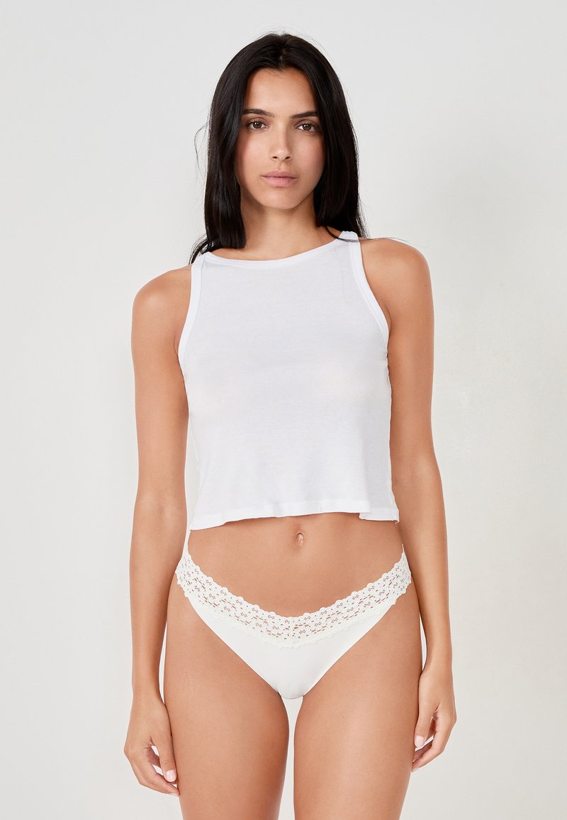 Weißes, kurzes Tanktop aus weichem Stoff mit rundem Halsausschnitt. Kombiniert mit weißer Unterwäsche, die einen Spitzenbesatz am Bund hat.