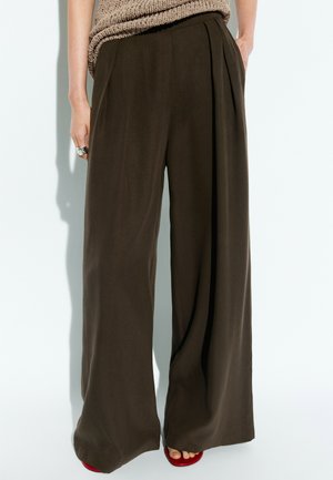Donna che indossa pantaloni scuri a gamba larga con un top beige lavorato a maglia, in piedi con le mani in tasca e scarpe rosse aperte in punta visibili.