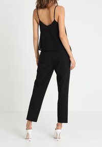 Haut sans manches noir avec de fines bretelles, associé à un pantalon noir ajusté. Le mannequin porte des chaussures à talons blancs. Design simple et épuré.