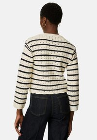 Sweter z krótkim rękawem w kremowym kolorze z czarnymi paskami i teksturowanym wzorem, posiadający okrągły dekolt i prążkowany dół, zestawiony z ciemnymi dżinsami.