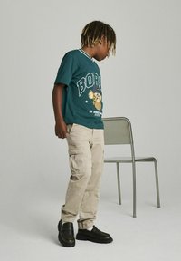 Camiseta verde con la impresión "BORN" y un gráfico de dibujos animados, pantalones cargo beige y zapatos negros sin cordones. Fondo sencillo con una silla.