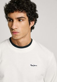 Sudadera blanca con un acabado texturizado, cuello redondo y un acento azul oscuro en la parte inferior. Presenta el logo de "Pepe Jeans" en náutico en el pecho.