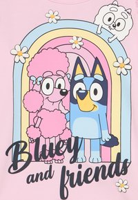 Jasnoróżowa koszulka z postaciami z kreskówki: niebieski pies z żółtymi akcentami i różowy pudel na tle tęczy, ozdobiona stokrotkami.