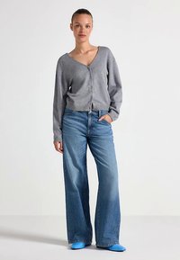 Cardigã cinzento de malha com decote em V, fecho de botões, combinado com jeans de perna larga em denim. Sabrinas azul vivo completam o visual.