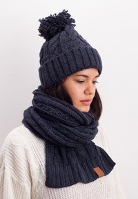 Dark gray knitted beanie with a pom-pom and matching scarf, featuring ribbed texture and a leather tag detail. Soft, chunky design.