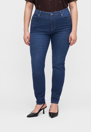 Woman wearing dark blue slim-fit jeans and black pointed-toe heels, standing against a plain light background.