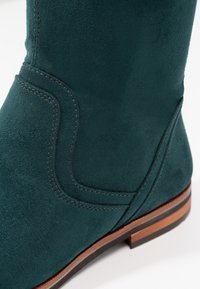 Grøn suedestøvle med en buet sømdetalje og en lav stablet hæl med et træelement. Glat tekstur og rund form.