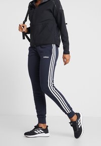 Svart zip-up jacka med en struktur med grå krage, ihop med marinblå joggers som har tre vita ränder längs sidorna, bärs med svarta sneakers.