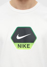 Εξάγωνο λογότυπο της Nike με λευκή γραμμή Swoosh και μαύρο κείμενο "NIKE" σε πράσινο φόντο στο μπροστινό μέρος ενός λευκού T-shirt.