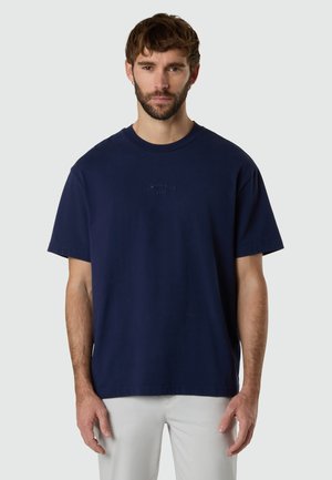 Mann mit kurzem braunem Haar und Bart, der ein dunkelblaues Kurzarm-T-Shirt mit dem Logo "North Sails 1957" und weiße Hosen trägt.