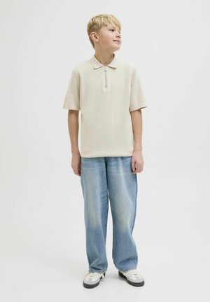 Hellbeige Polohemd mit kurzen Ärmeln und einem Frontreißverschluss, kombiniert mit lockeren, hellblauen Jeans und weißen Sneakers.