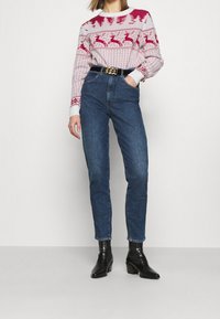 Rot-weiß gemusterter Pullover mit Rentieren und Bäumen, kombiniert mit hochgeschnittenen blauen Jeans und schwarzen Absatzstiefeletten.