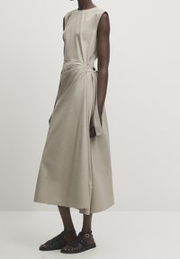 Robe portefeuille sans manches beige, fixée par un bouton sur le côté et un lien, associée à des sandales plates à brides noires sur une personne debout.