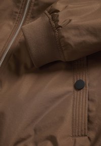 Chaqueta bomber marrón con tela suave, puños acanalados y cremallera metálica. Incluye un detalle de botón a presión negro en el lateral.