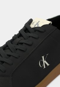 Zwarte sneaker met gestructureerde synthetische bovenkant, platte veters en een wit logo op de tong. Gumrubberen zool met een zichtbaar profiel.