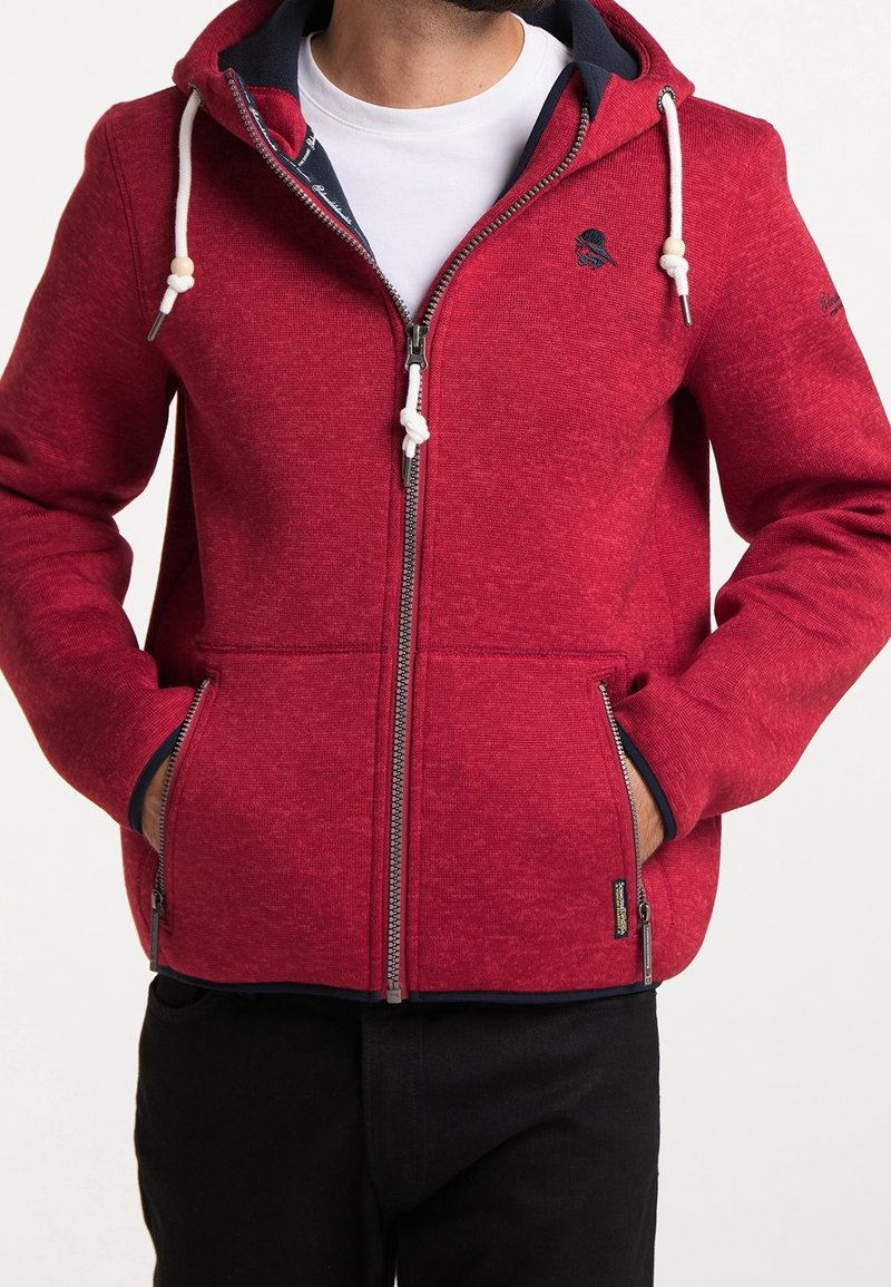 Punainen vetoketjullinen huppari, valmistettu teksturoidusta kankaasta, jossa on kiristysnyöri huppua varten, etutaskut ja tummansininen kontrastivärinen sisävuori.