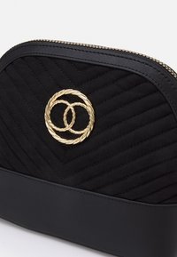 Černá prošívaná taška z látky s hladkým černým dnem, obsahující zlaté logo se spletitým kruhem a zlatý zip.