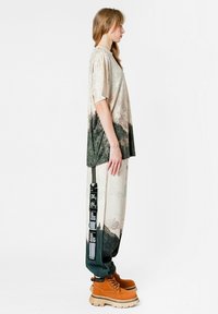 Camiseta texturizada XL en beige y verde con diseño abstracto, combinada con pantalones holgados de estampado que tienen una correa negra y botas naranjas.